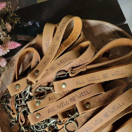 A collection of tan leather wrist straps with metal clasps and engraved text reading 'ALL WILL BE WELL.' arranged in a wooden bowl on a dark wooden table.